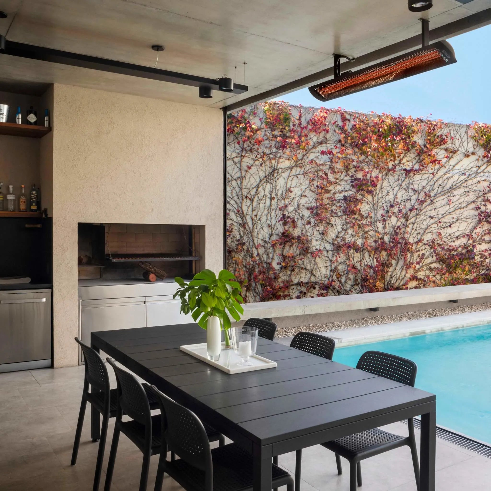 Outdoor dining area beside a pool, featuring a ceiling-mounted Bromic Tungsten Electric Heater, warming a stylish patio with modern furnishings and natural accents.