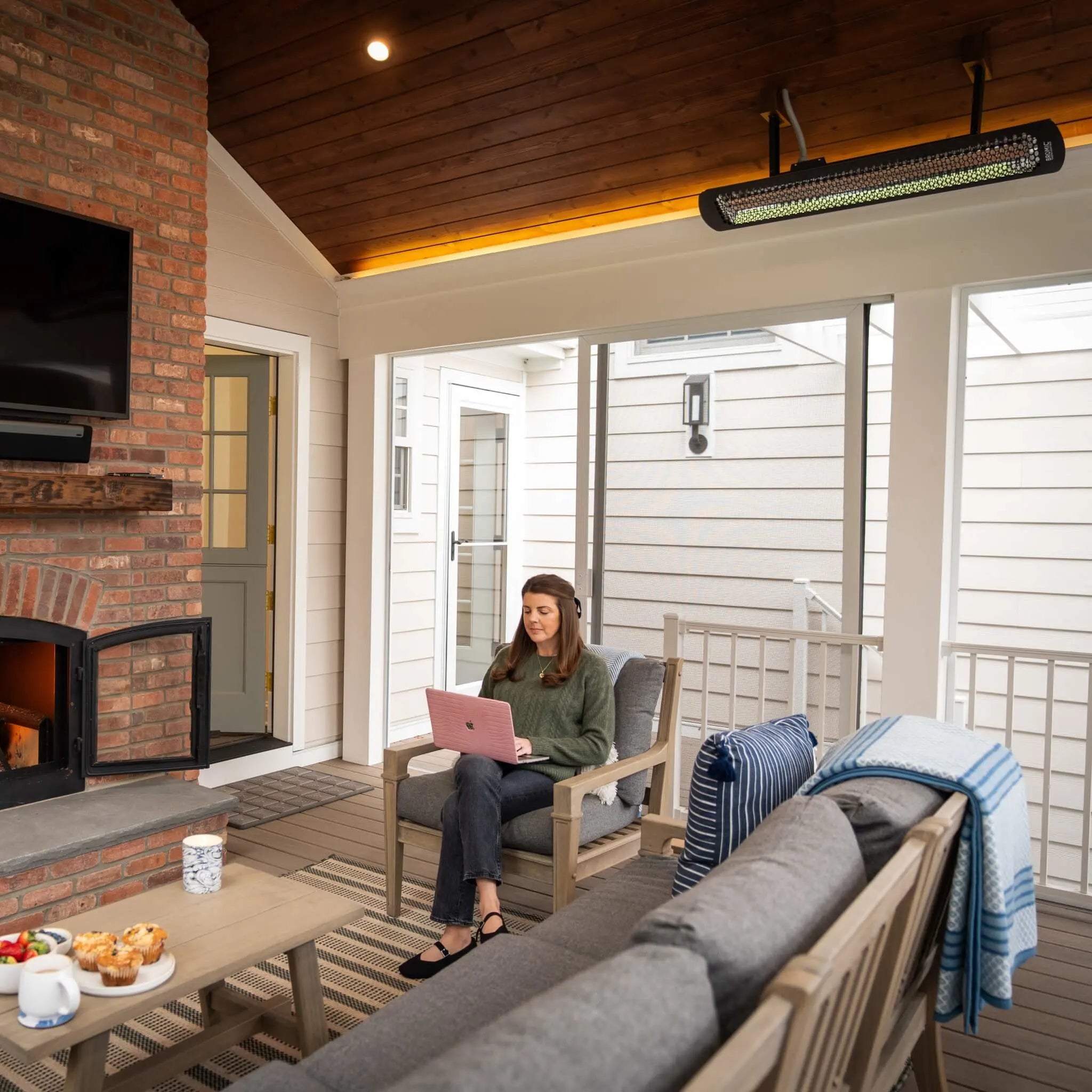 Enclosed patio space with a Bromic Heating Tungsten Smart-Heat Electric Heater installed overhead, providing warmth as a woman works on her laptop beside a lit fireplace.