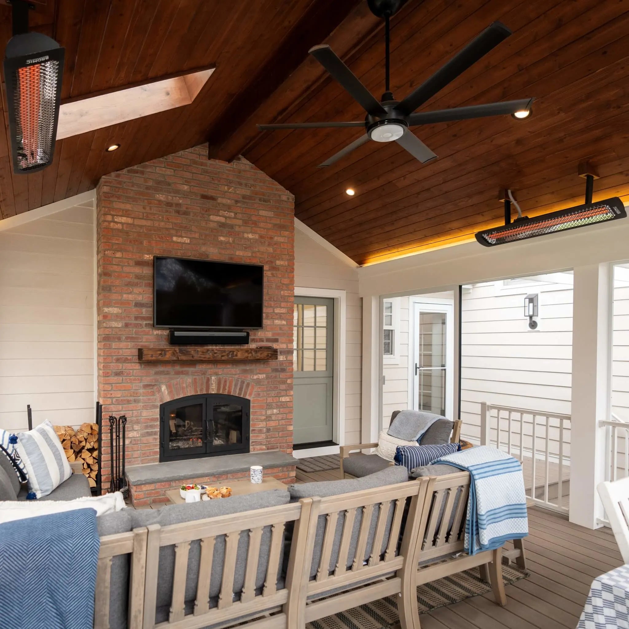 Spacious enclosed patio with two Bromic Tungsten Smart-Heat Electric Heaters on a wood-paneled ceiling, providing radiant warmth for a cozy family living area with fireplace and ceiling fan.