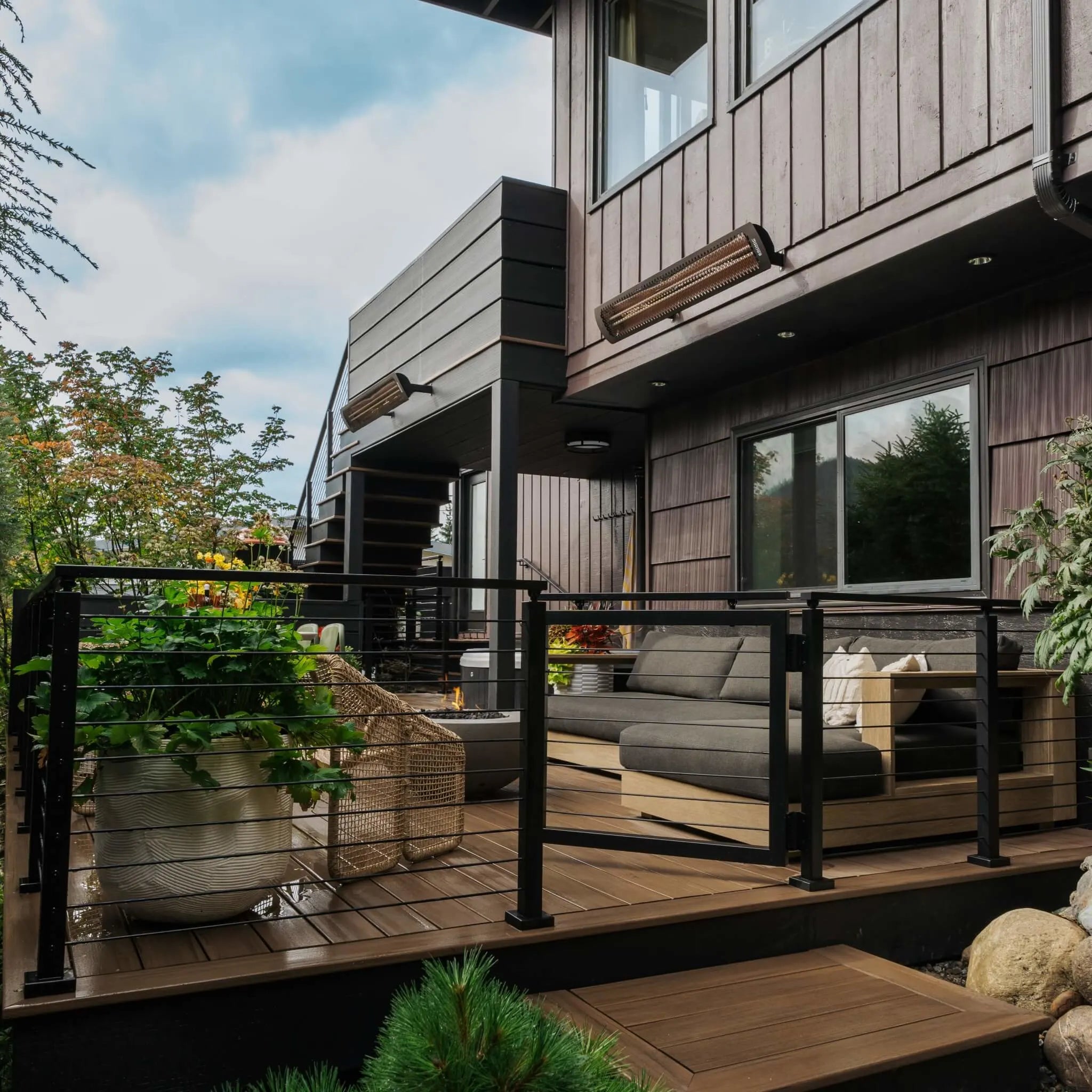 Modern outdoor deck with Bromic Tungsten Electric Smart-Heat heaters mounted along a dark wood exterior wall, warming a cozy patio lounge area with sleek furniture and lush greenery.