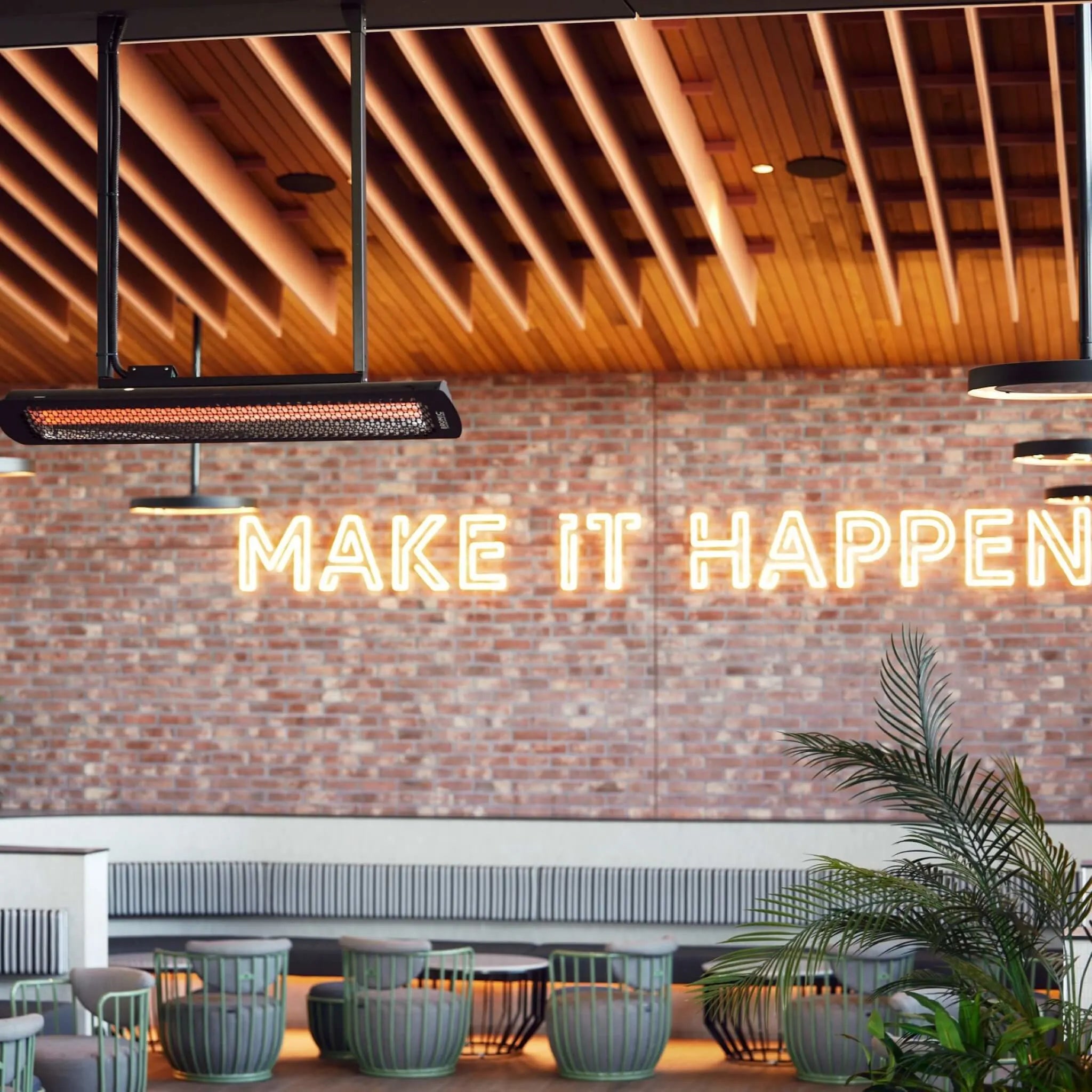 Contemporary restaurant interior featuring Bromic Tungsten Electric Smart-Heat heaters ceiling-mounted above a â€œMAKE IT HAPPENâ€ neon sign, blending warmth and industrial style for comfortable dining.