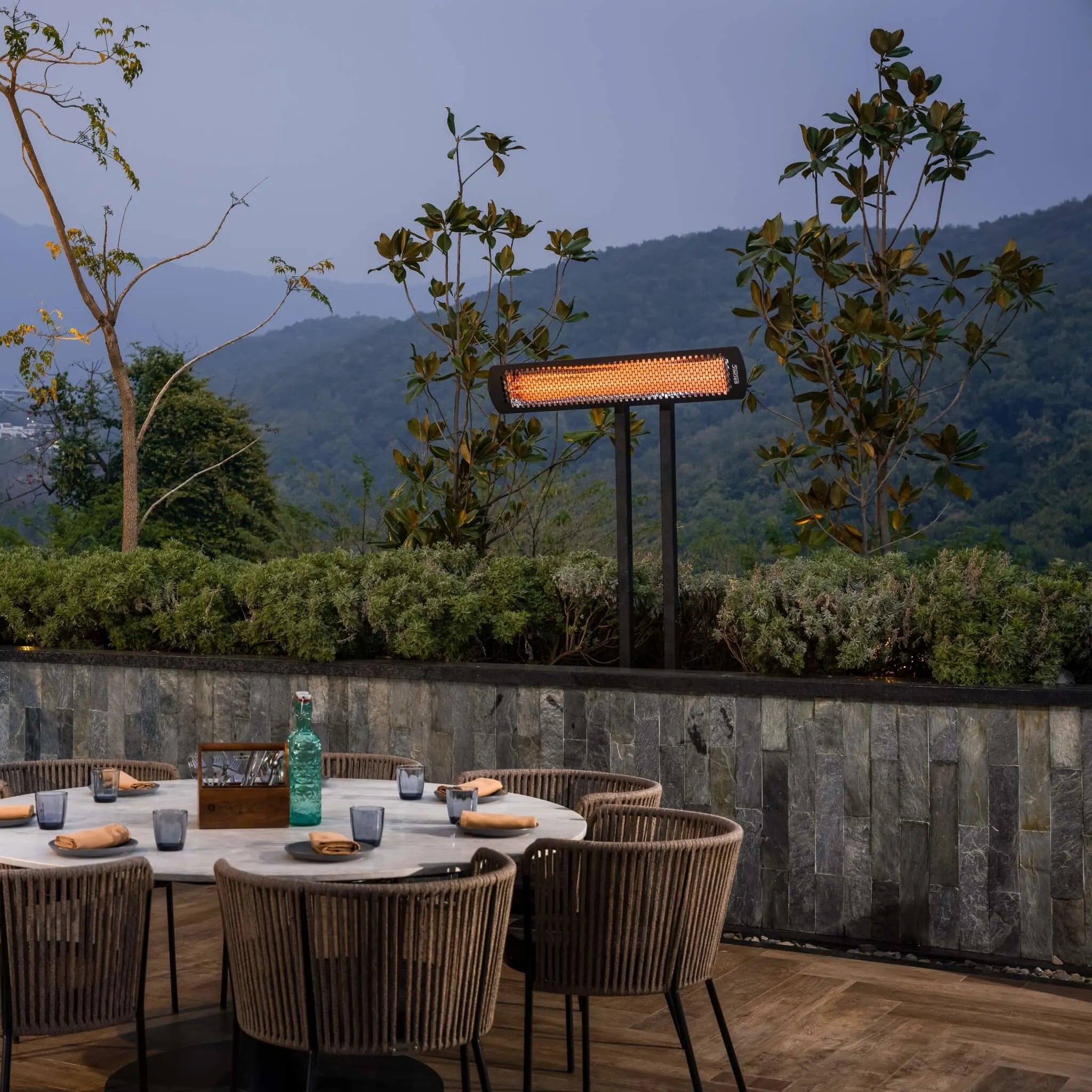 Elegant outdoor restaurant terrace with Bromic Tungsten Electric Smart-Heat heater on a post mount, providing gentle warmth over a dining table surrounded by mountain views.