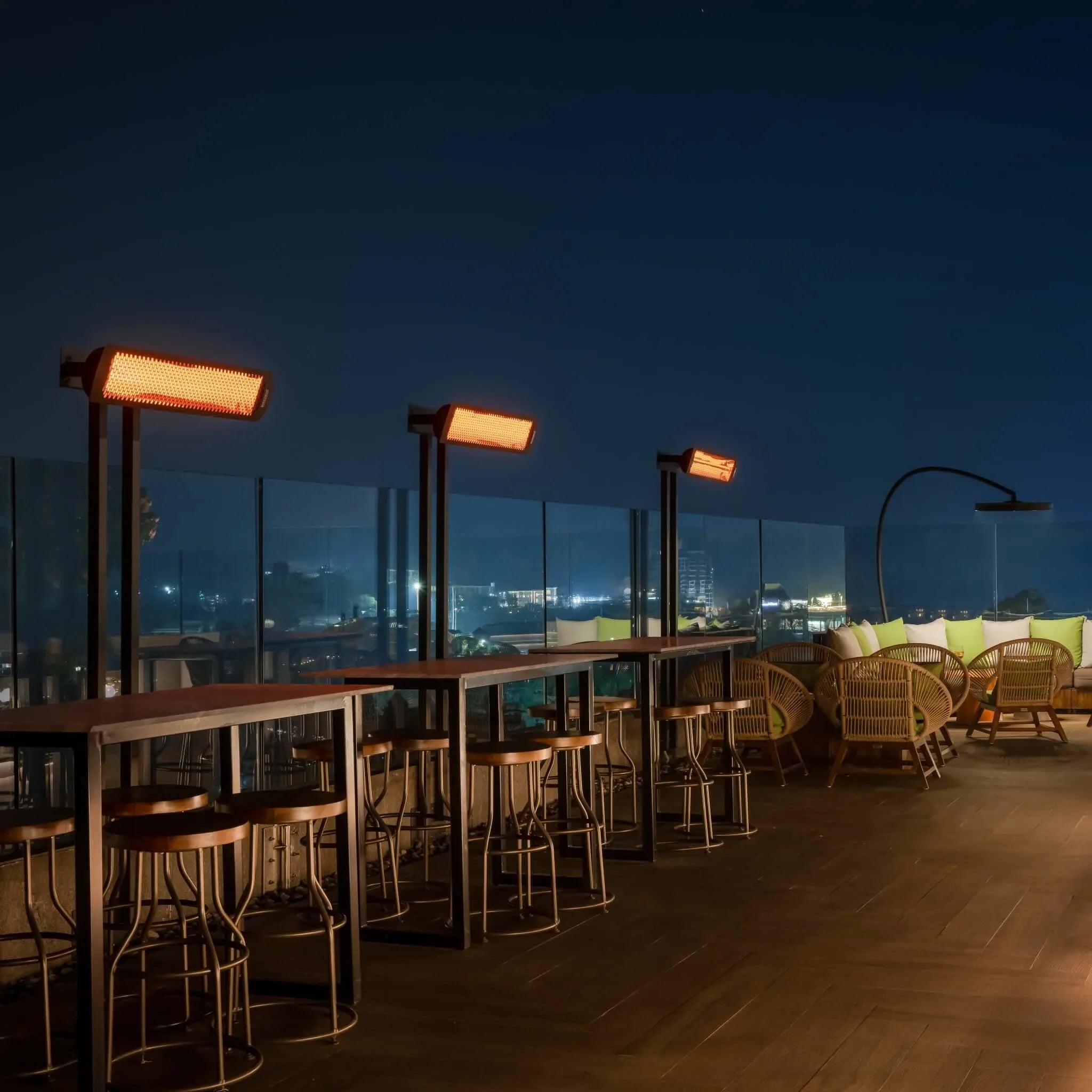 Nighttime rooftop bar with several Bromic Tungsten Electric Smart-Heat heaters, creating a comfortable outdoor experience with warm amber lighting and scenic skyline views.