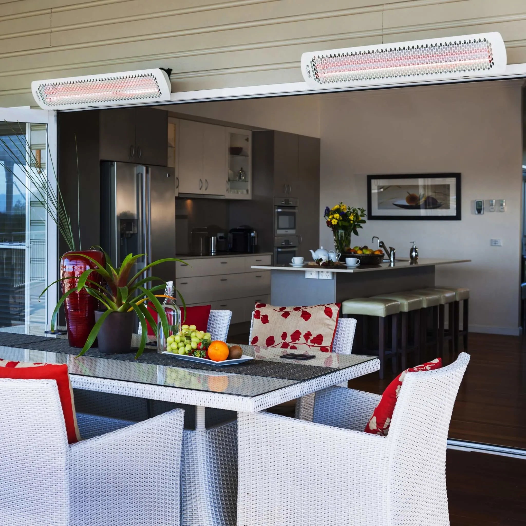 Elegant outdoor dining space blending indoor and outdoor living, featuring white Bromic Tungsten Electric Smart-Heat heaters above a wicker table set with bright red and white dÃ©cor.