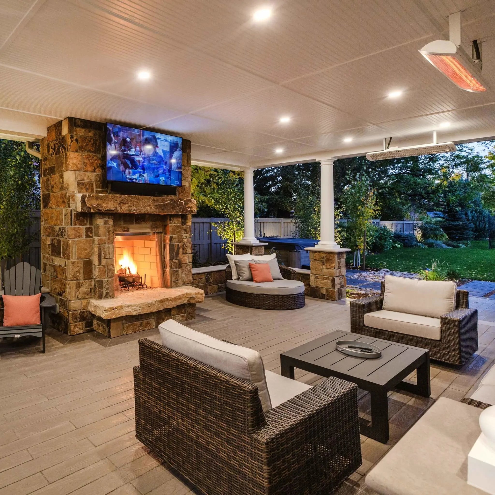 Inviting backyard patio with Bromic Tungsten Electric Smart-Heat heaters installed overhead, illuminating a wicker furniture set beside a roaring stone fireplace and lush garden view.