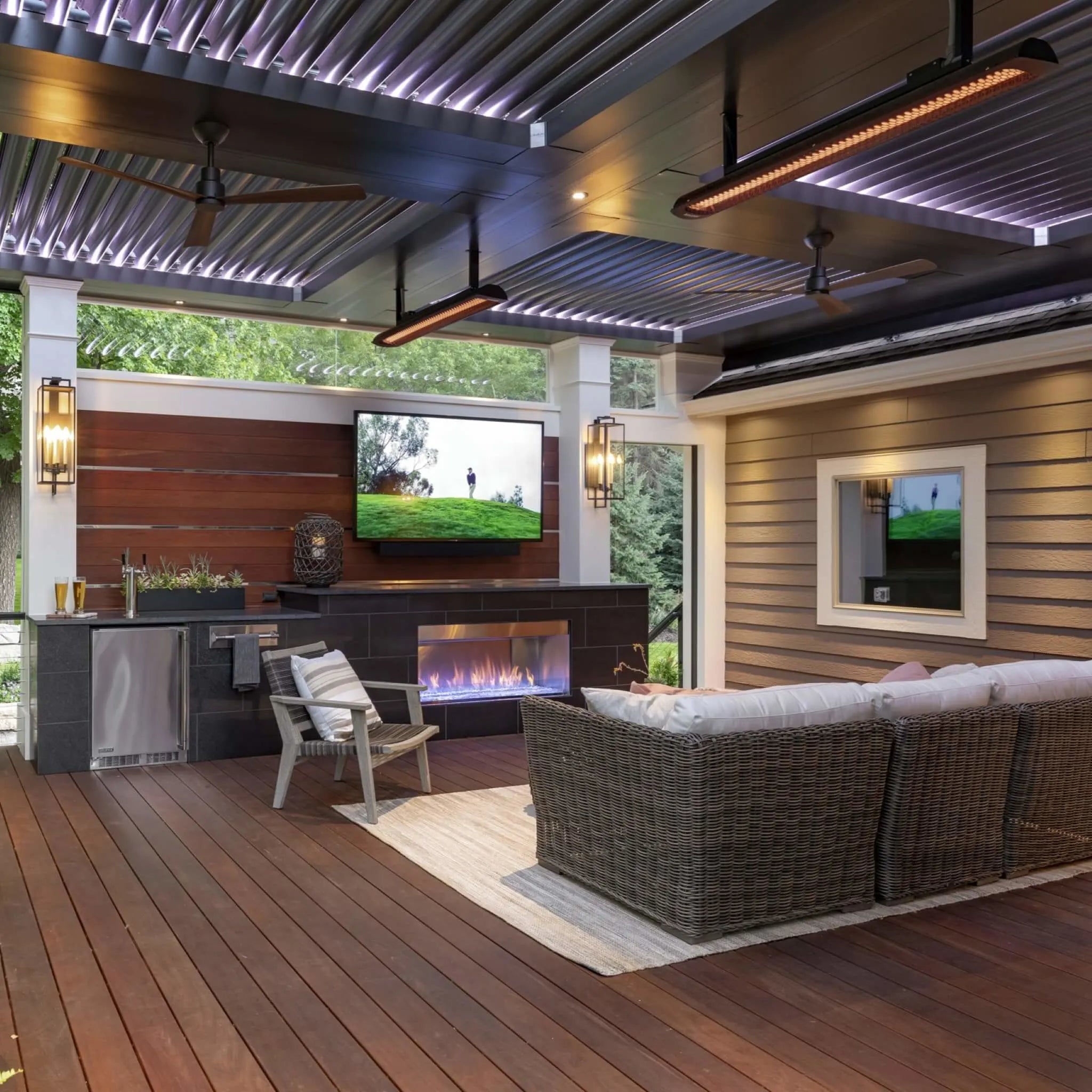 Elegant outdoor living area featuring Bromic Tungsten Electric Smart-Heat heaters on a modern covered patio with wicker furniture, a built-in TV, and a sleek gas fireplace.