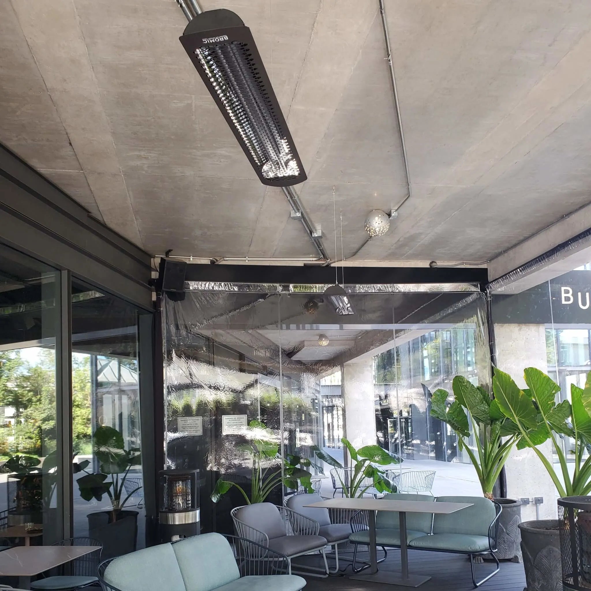 Modern cafÃ© in Chile with ceiling-mounted Bromic Tungsten Electric Smart-Heat heaters, providing radiant warmth in a semi-enclosed patio surrounded by greenery and glass walls.
