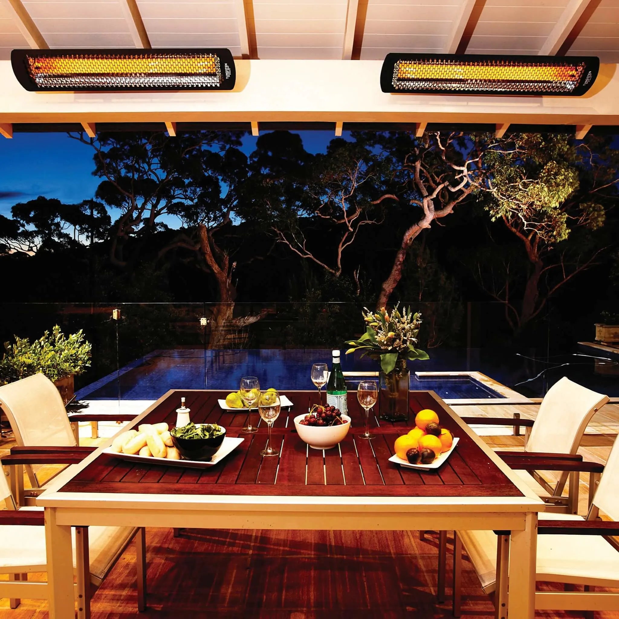 Elegant covered patio featuring dual Bromic Heating Tungsten Electric Smart-Heat heaters illuminating a warm evening dining setup beside a pool, surrounded by lush trees and ambient lighting.