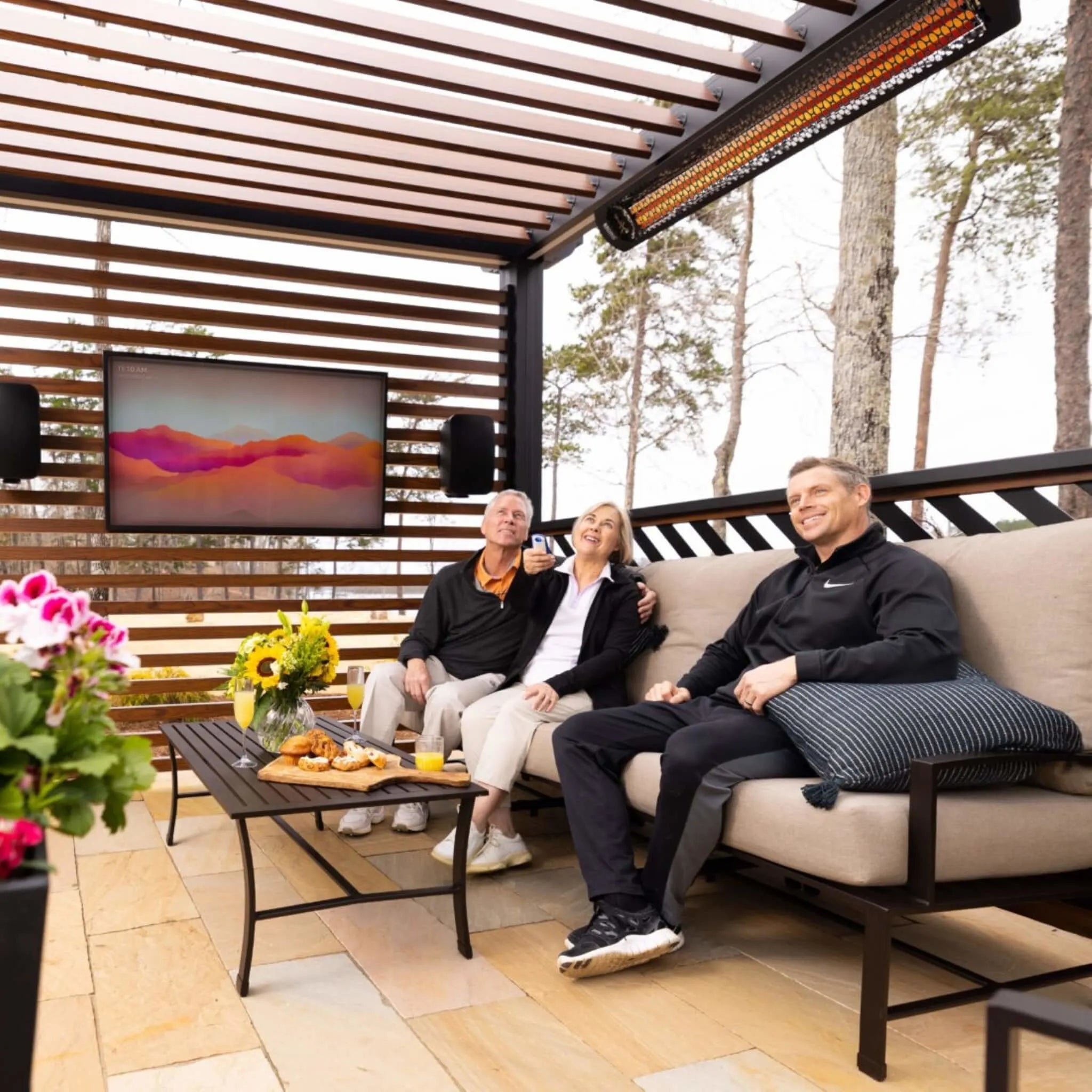 Outdoor entertainment space with family relaxing under a slatted pergola, warmed by Bromic Tungsten Electric Smart-Heat heaters while enjoying drinks and a wall-mounted television.