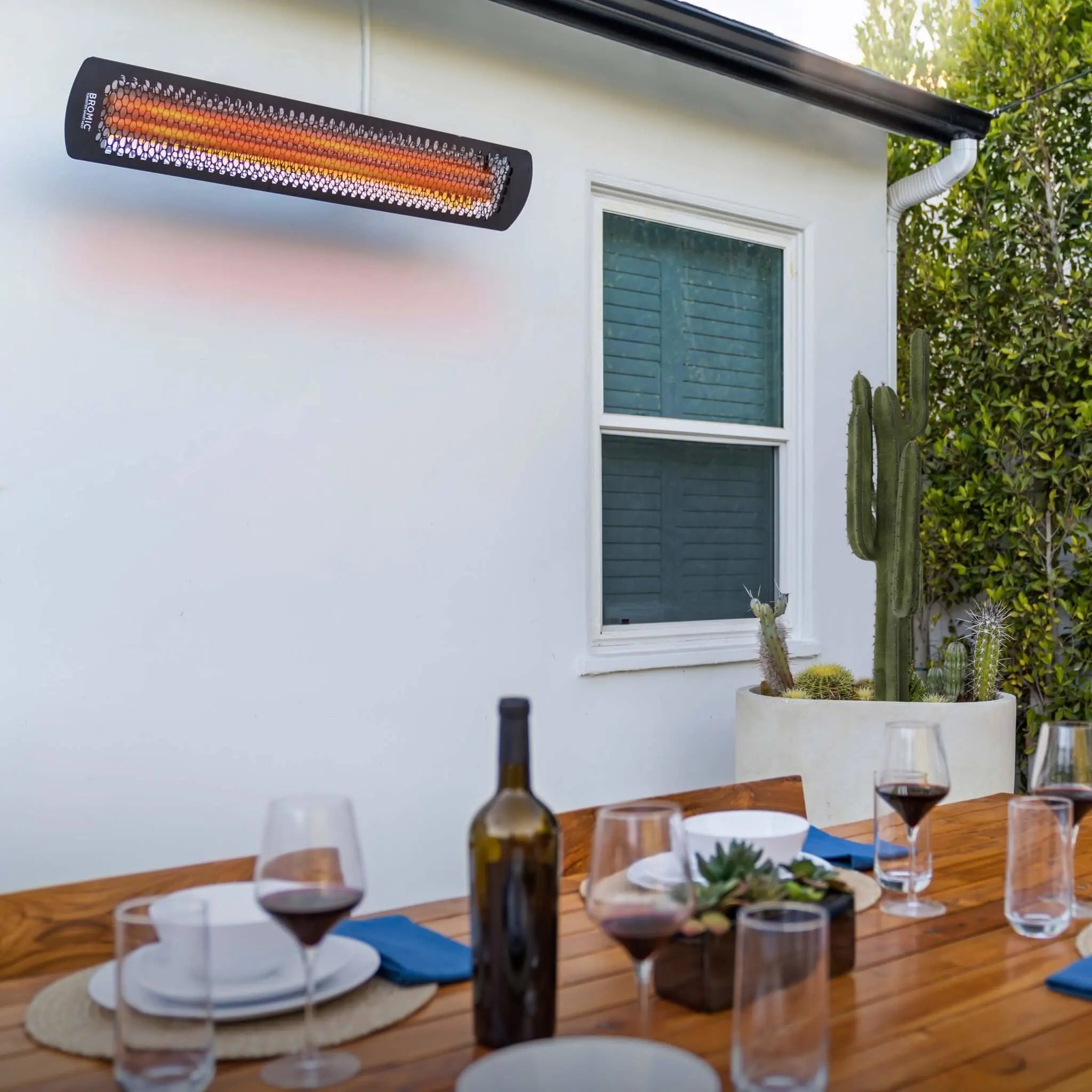 Modern outdoor dining setup with a Bromic Tungsten Electric Smart-Heat heater mounted to a white wall, providing radiant heat during a relaxed wine and dinner evening.