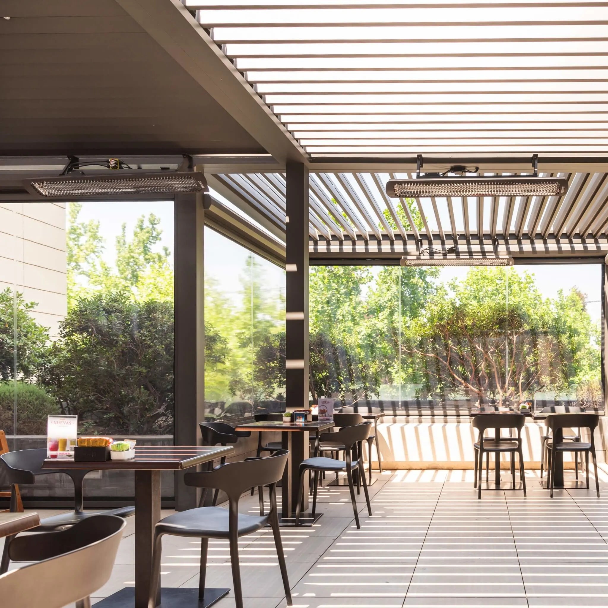 Modern cafÃ© patio with Bromic Heating Tungsten Electric Smart-Heat heaters mounted beneath a slatted pergola roof, providing comfortable radiant warmth for outdoor dining guests.