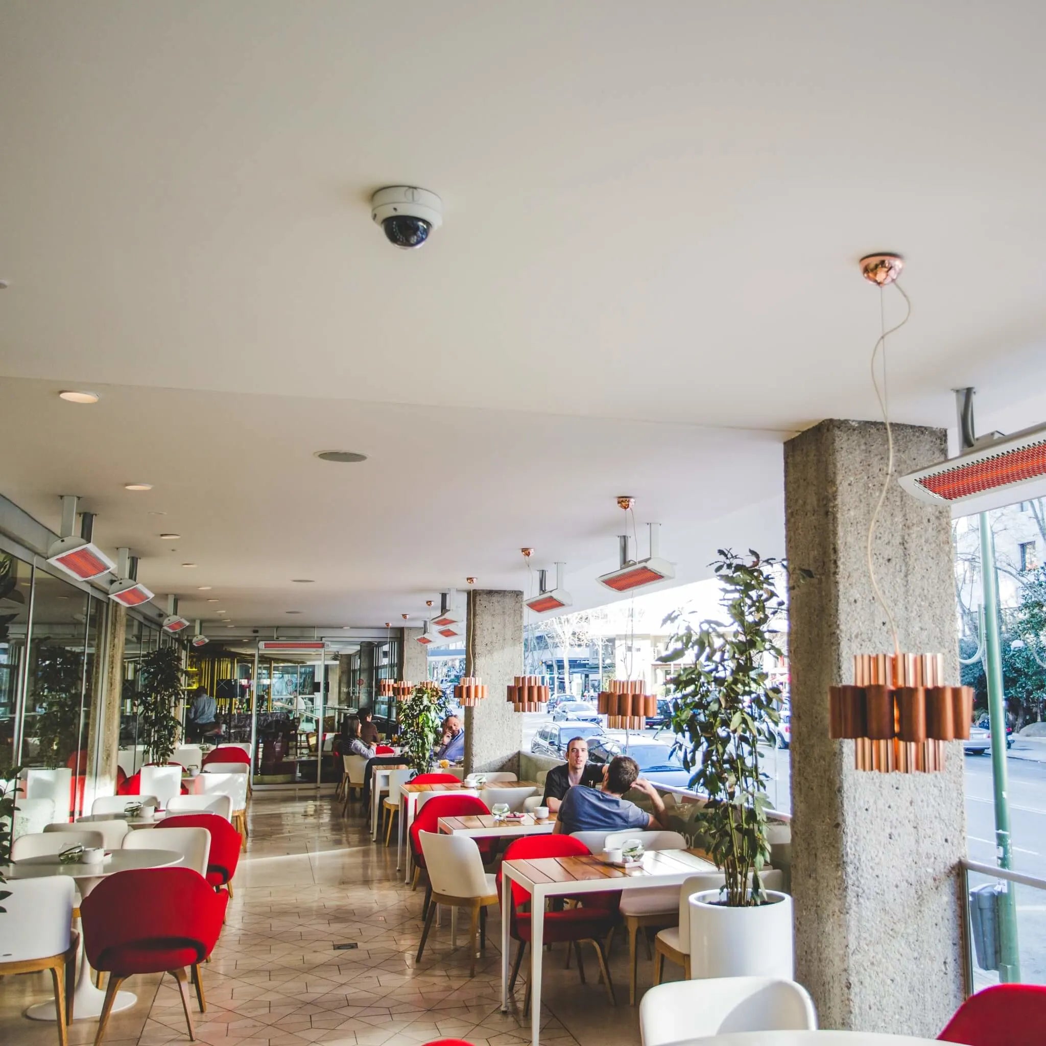 Bright cafÃ© interior with Bromic Heating Tungsten Electric Smart-Heat white heaters mounted along the ceiling, keeping guests comfortably warm while dining at modern red and white tables.